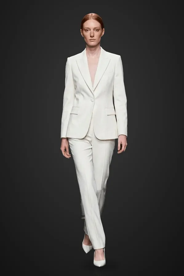 Modelo pelirroja con traje blanco de chaqueta y pantalón camina como en un desfile, sobre fondo oscuro